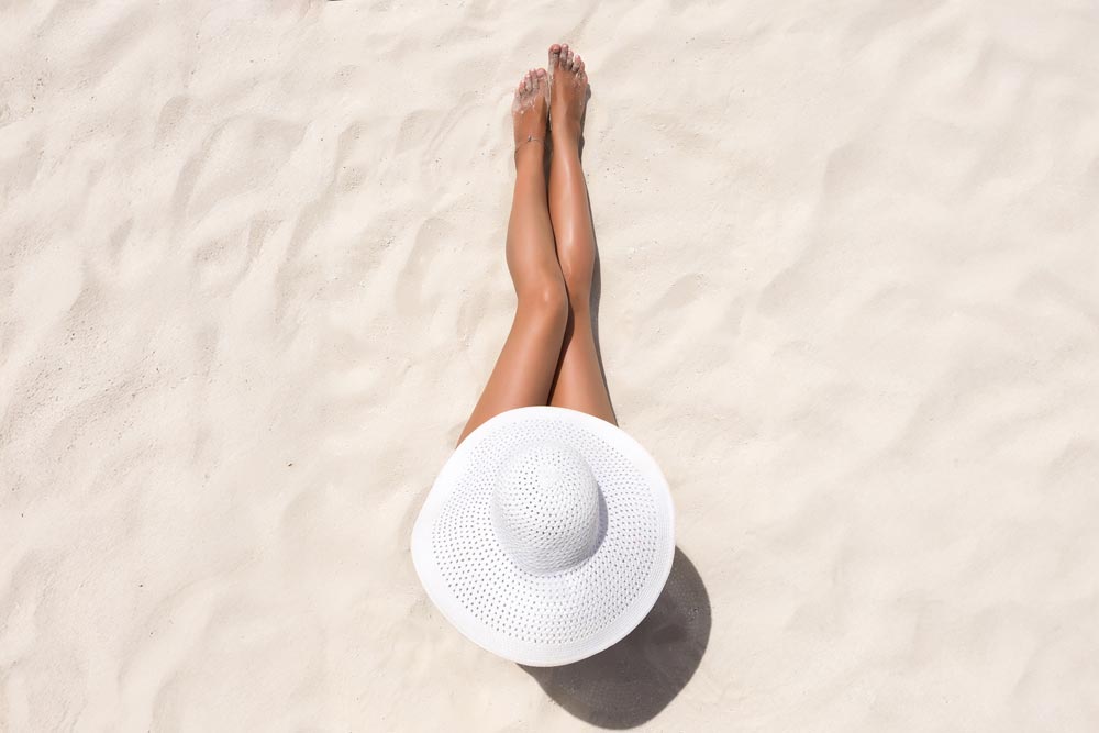 En somrig strandbild tagen från ovanifrån som visar en kvinna liggande på ljus sand med benen sträckta uppåt.